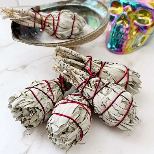 Bundles of Organic White Sage Torch Smudge Stick, tied with red string, are arranged on a white marble surface. A metallic rainbow-colored skull and a shiny shell dish add decorative elements to this cleansing and purifying scene.