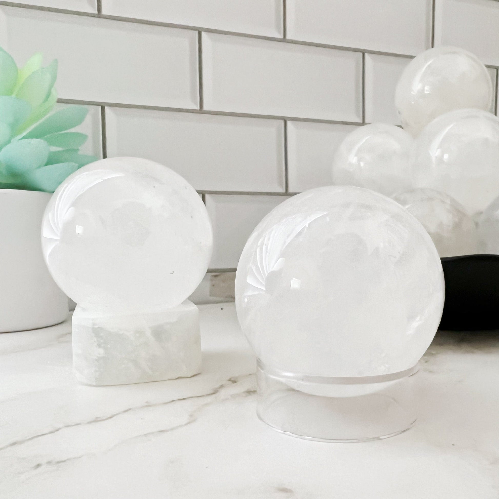 The image shows several 40mm Clear Quartz Crystal Balls of varying sizes placed on a white counter with a white subway tile backsplash. One healing crystal rests on a square white marble base, and there is a green succulent plant in the background.