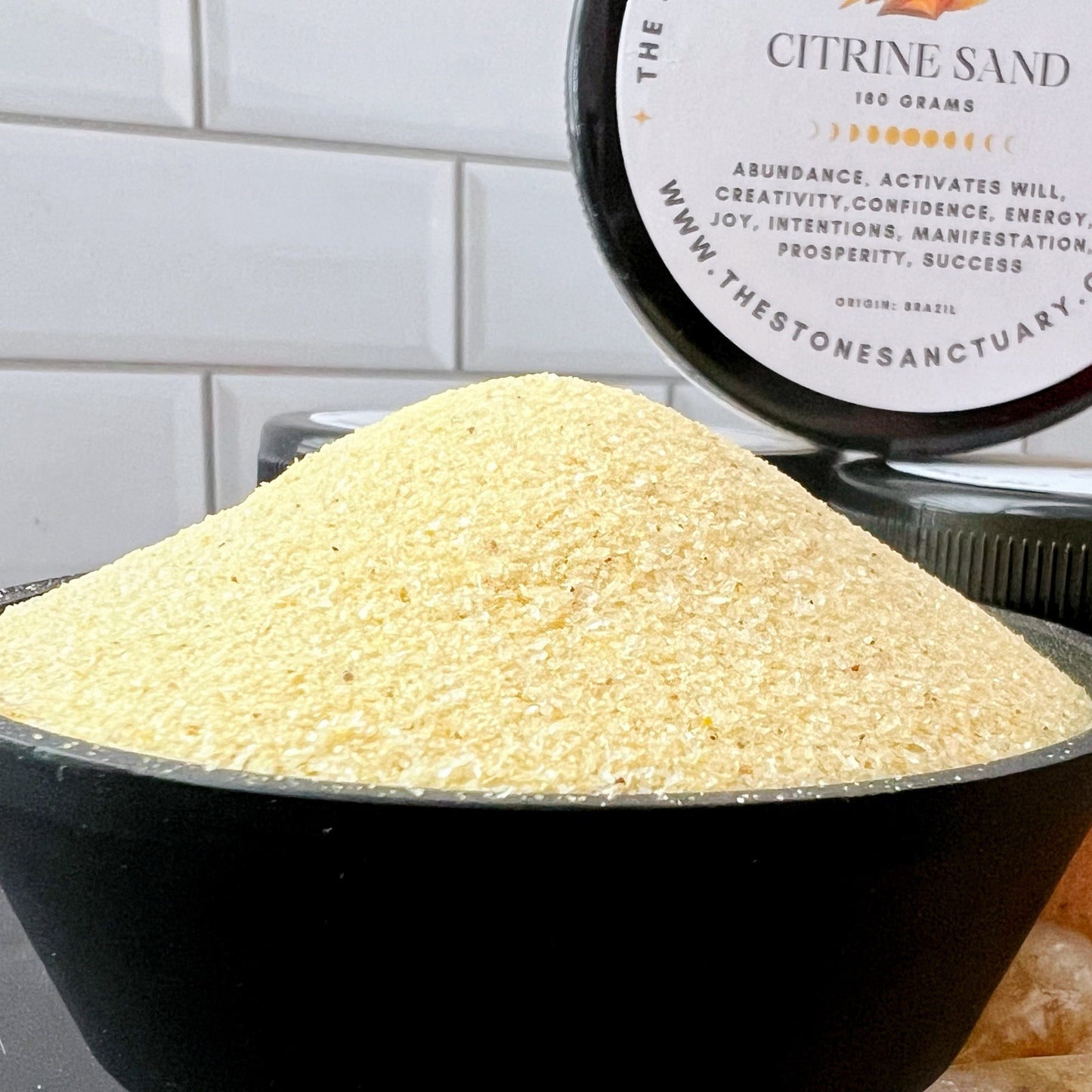 A small black bowl filled with Citrine Gemstone Sand sits beside an open jar and a closed jar of the same sand labeled “Citrine Gemstone Sand - 100 grams.” Four polished yellow stones, known for their healing properties and wealth attraction, are arranged in the foreground against a white tiled wall background.