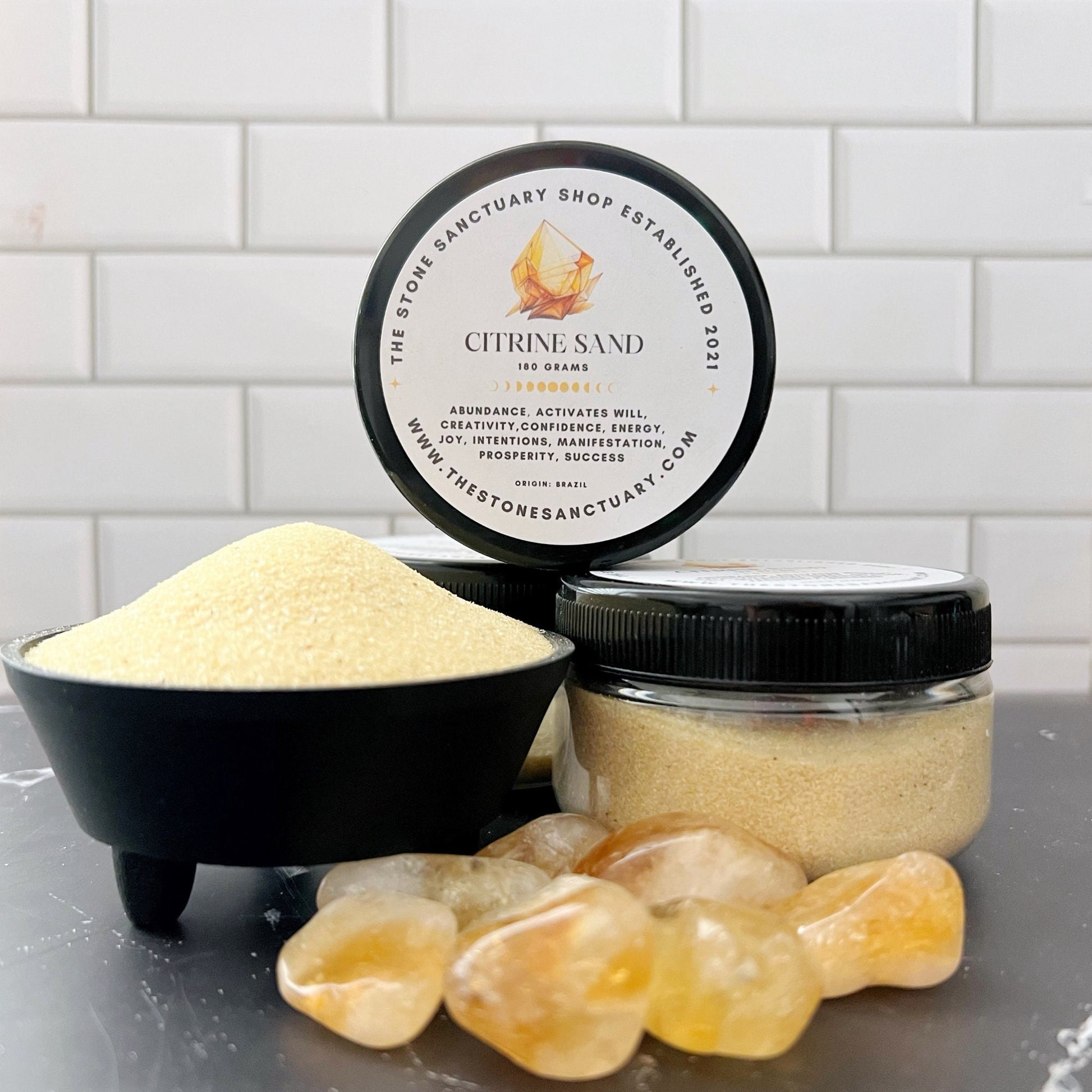 A small black bowl filled with Citrine Gemstone Sand sits beside an open jar and a closed jar of the same sand labeled “Citrine Gemstone Sand - 100 grams.” Four polished yellow stones, known for their healing properties and wealth attraction, are arranged in the foreground against a white tiled wall background.