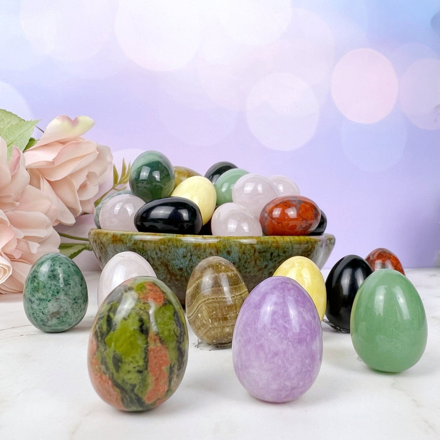 A collection of polished gemstone eggs, perfect for energy healing, is displayed on a light surface. Some 1" Mini Crystal Eggs are inside a greenish pottery bowl, while others are arranged around it. Pink roses are in the background, and the backdrop features soft bokeh lights.