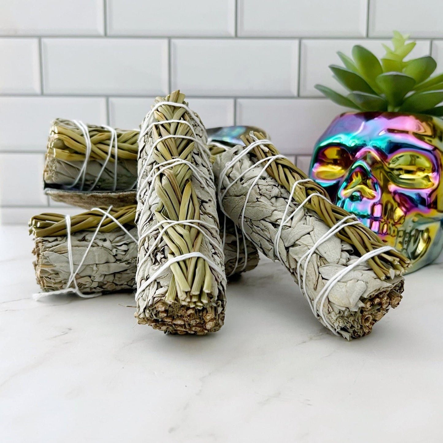 Bundles of **Organic White Sage with Sweetgrass** wrapped with white strings are arranged in front of a white tile background. A plant with green leaves sits inside a rainbow-colored skull-shaped planter in the background. The scene, perfect for cleansing negative energy, is set on a white marble surface.