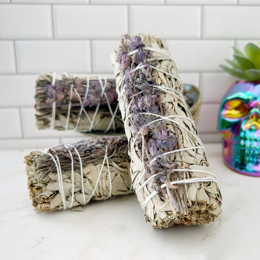 Three **Deluxe Organic White Sage & Lavender Smudge** sticks are bound with string and adorned with dried purple flowers. They rest on a white countertop against a tiled backsplash. In the background, a colorful skull ornament and a small potted succulent add charm to the serene scene.