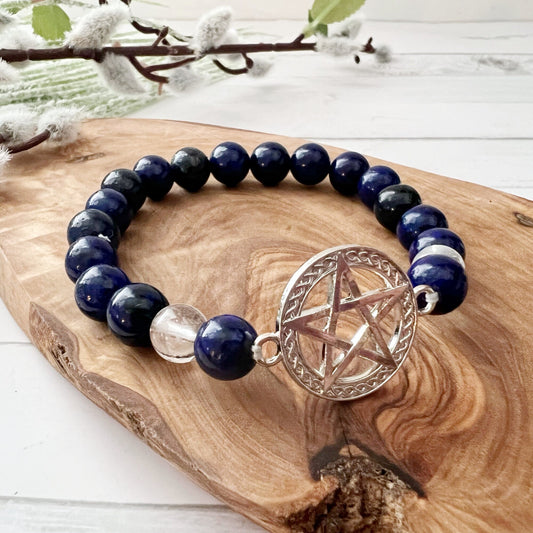 An 8mm Lapis Lazuli & Clear Quartz Beaded Bracelet with Pentacle Charm rests on a wooden surface. In the background, there’s a branch with soft, fuzzy buds and a textured green cloth, creating a rustic and natural setting that highlights this energy bracelet.