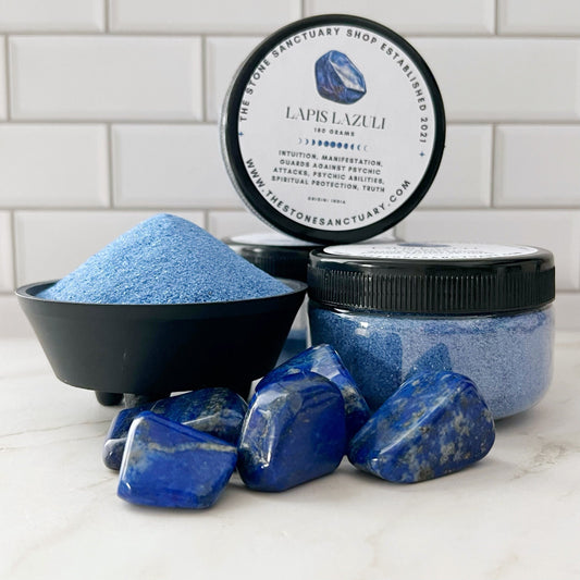 Three lapis lazuli stones and two jars of Lapis Lazuli Gemstone Sand are displayed. One jar has a lid that reads "The Stone Sanctuary Shop Established 2021" and "Lapis Lazuli Gemstone Sand." Known for their healing properties, these Lapis Lazuli crystals rest on a clean, white tiled background and marble surface, promoting inner peace.