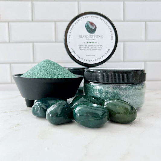 A display featuring a small round container labeled "Bloodstone Gemstone Sand," an open black jar filled with fine green powder, a closed jar of green gel, and a collection of polished green gemstones, all set against a white tiled background, radiating grounding energy.