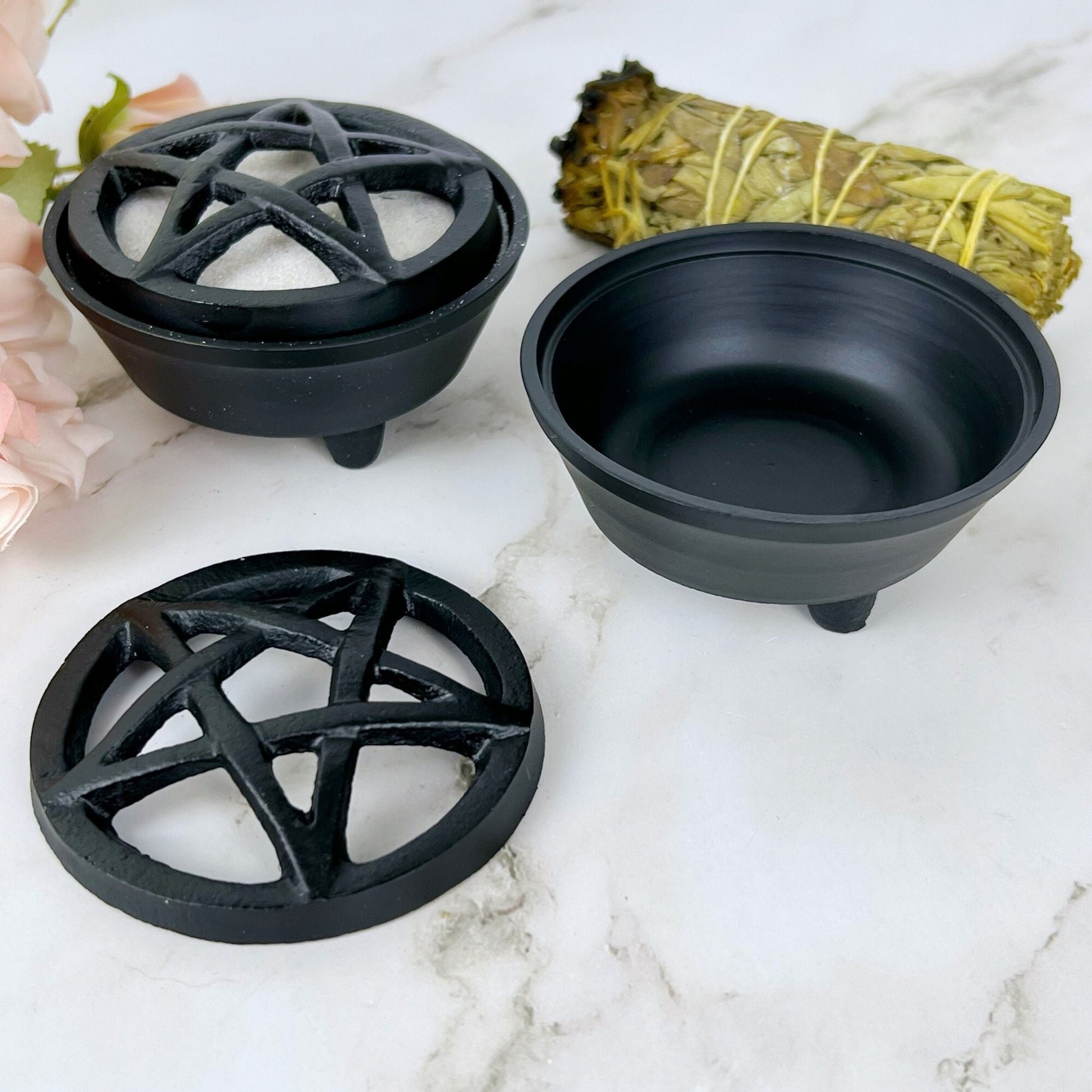 A Cast Iron Pentacle Incense Holder is displayed on a marble surface. The set is disassembled, showcasing the lid with the pentacle, the bowl, and a smudge stick wrapped in string. A few pink flowers are partially visible to the side.