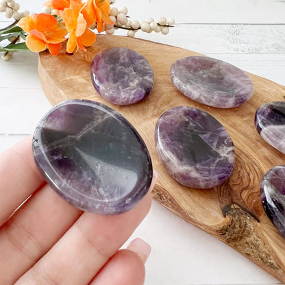 A hand holding a smooth, oval-shaped Amethyst Worry Stone, with several similar stones placed on a wooden board in the background. An orange flower bouquet lies on the top left corner. The setup is on a white wooden surface, ideal for those seeking psychic protection or to relieve stress.