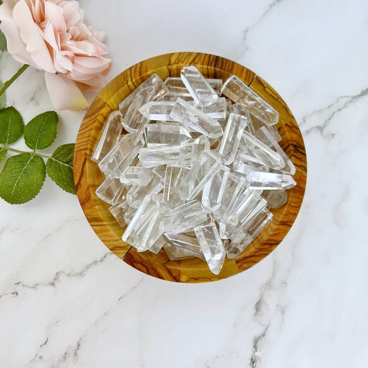 A wooden bowl filled with multiple 1-2" Mini Clear Quartz Points, known as the master healer, sits on a marble surface. A pale pink rose with green leaves lies next to the bowl, adding a touch of softness to the natural and serene composition.