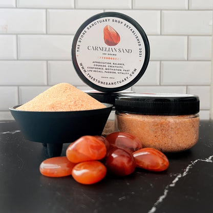 A jar filled with Carnelian Gemstone Sand and accompanied by a small pile of the sand in a black dish. Several polished carnelian stones are placed in front of the jar and dish. The jar's label includes details about the Carnelian Gemstone Sand, its healing properties, and its various uses for balancing the sacral chakra.