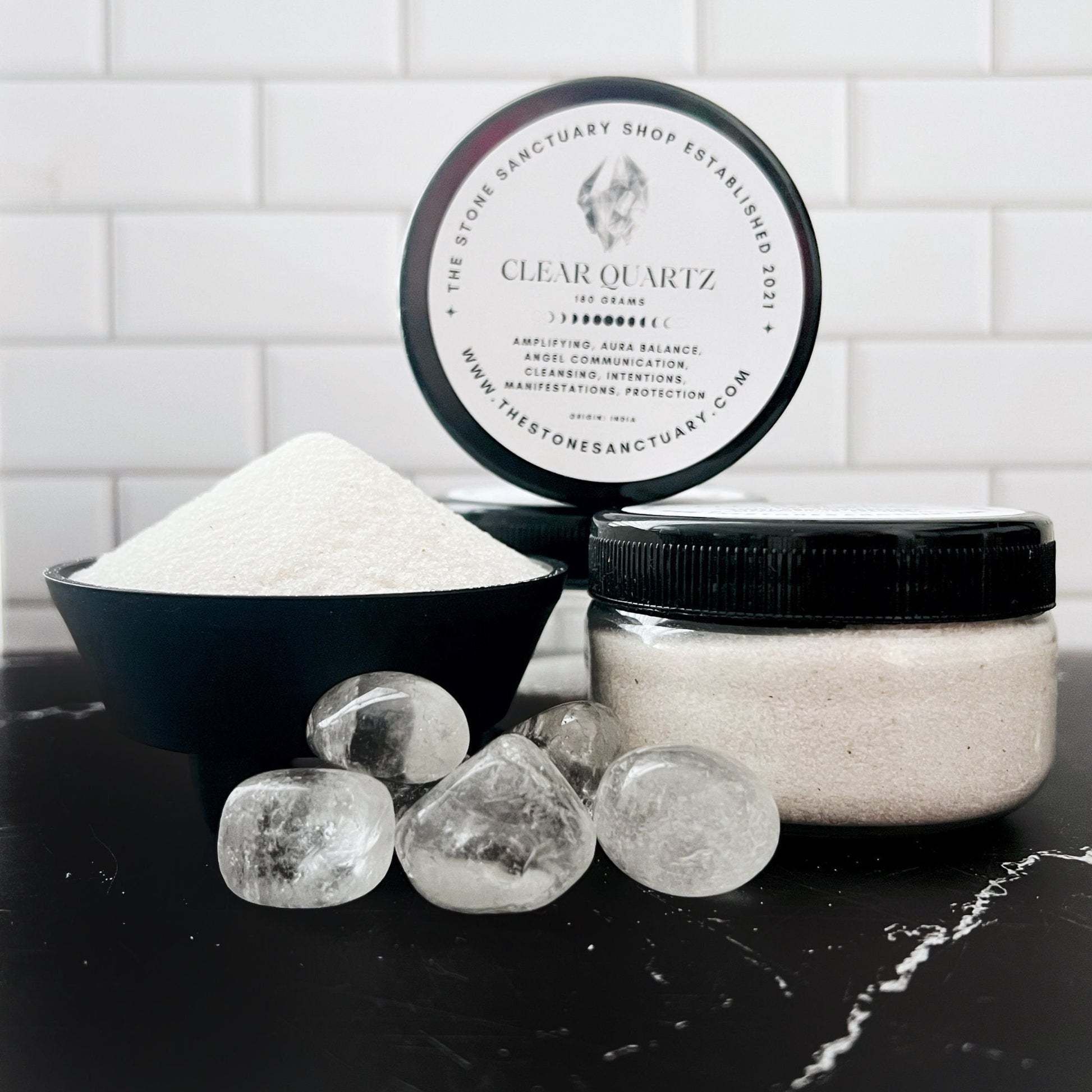 A small open jar labeled "Clear Quartz Sand" filled with white powder, likely Quartz Crystal Sand, is placed beside a black bowl also containing white powder. Several natural quartz crystals are scattered in front of the items. A lid with text is leaned against the jar. The background is a white subway tile wall.