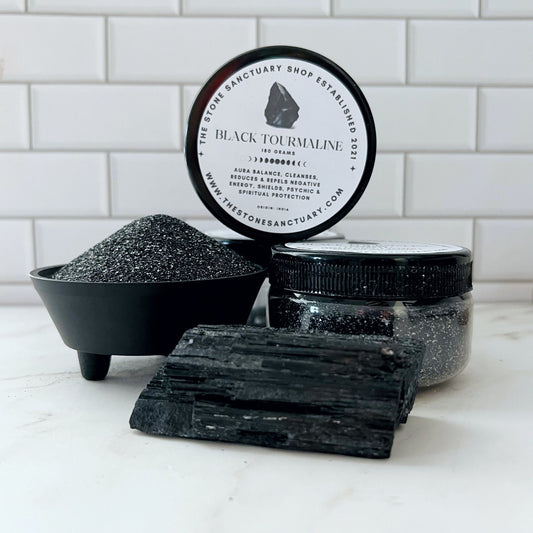 A small black bowl filled with Black Tourmaline Gemstone Sand sits on a white surface. Next to it, an open jar of gemstone sand and a raw black tourmaline crystal are displayed in front of a white tiled background, showcasing the protective properties these natural elements offer.