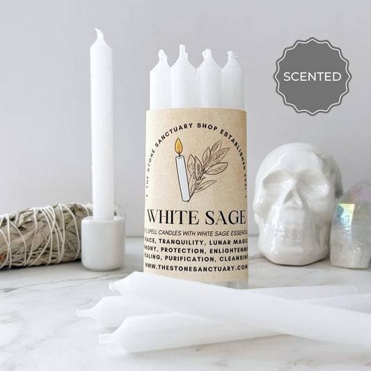 A cylindrical pack of 5" White Sage Scented White Chime Candles labeled "White Sage" and "The Stone Sanctuary Shop." The pack, perfect for purification or spellwork, is surrounded by individual white candles, a white skull decoration, a bundle of sage, and a crystal. A "Scented" label is shown on the edge.