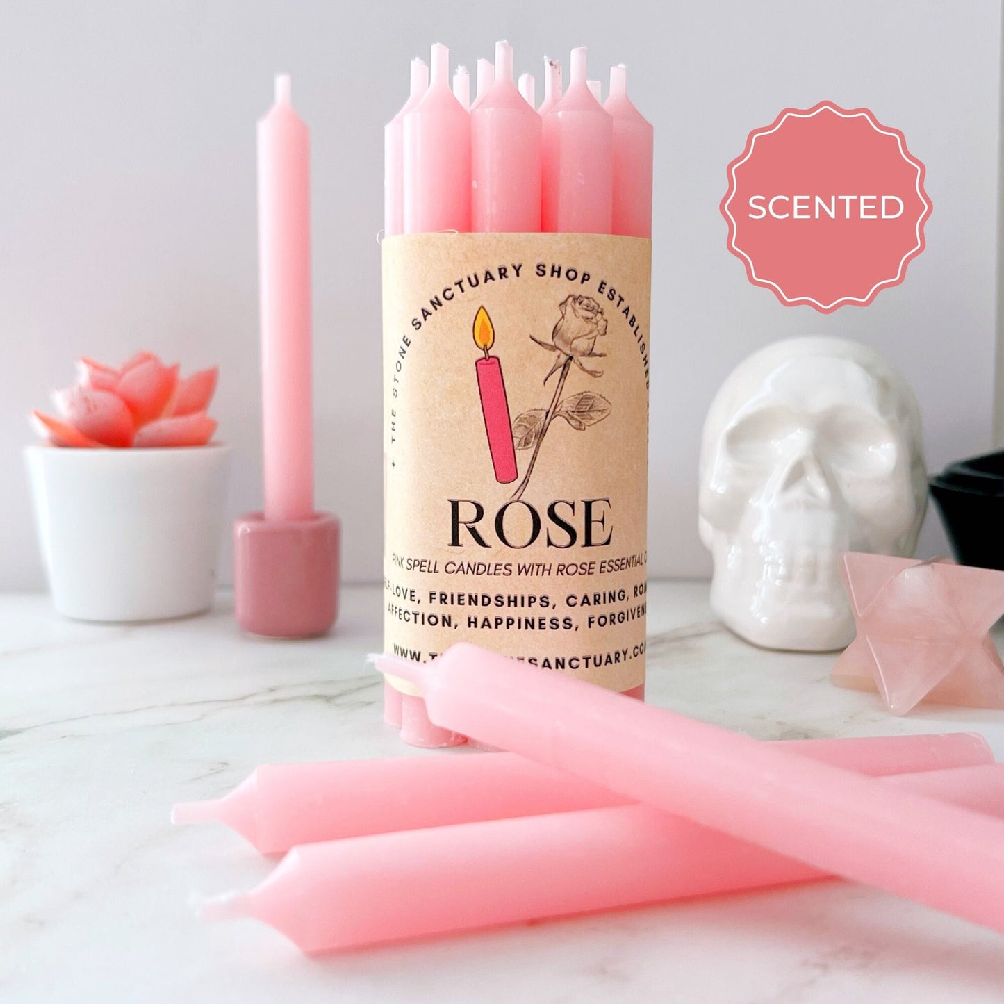 A group of 5" Rose Scented Pink Chime Candles labeled "ROSE" is displayed in a cozy setting. The candle packaging features a rose illustration. Nearby, a small pink succulent, a white skull, and a pink crystal contribute to the serene ambiance. A label indicates the candles are scented with rose essential oil, perfect for spellwork.