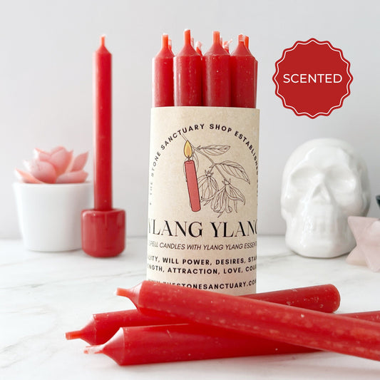 A cluster of 5" Ylang Ylang Red Chime Candles is displayed on a white surface. The candles are wrapped with a label featuring a red candle illustration and floral designs. In the background are a ceramic skull, a small pink flower, and other red candles.