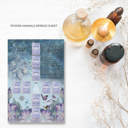 A card deck titled "Spirit Of The Animals Oracle" by Jody Bergsma is laid on a white wooden surface. The cover features a dragonfly and water lilies, embodying the essence of Animal Spirit Cards. Surrounding the deck are purple flowers and small purple petals scattered on the surface.