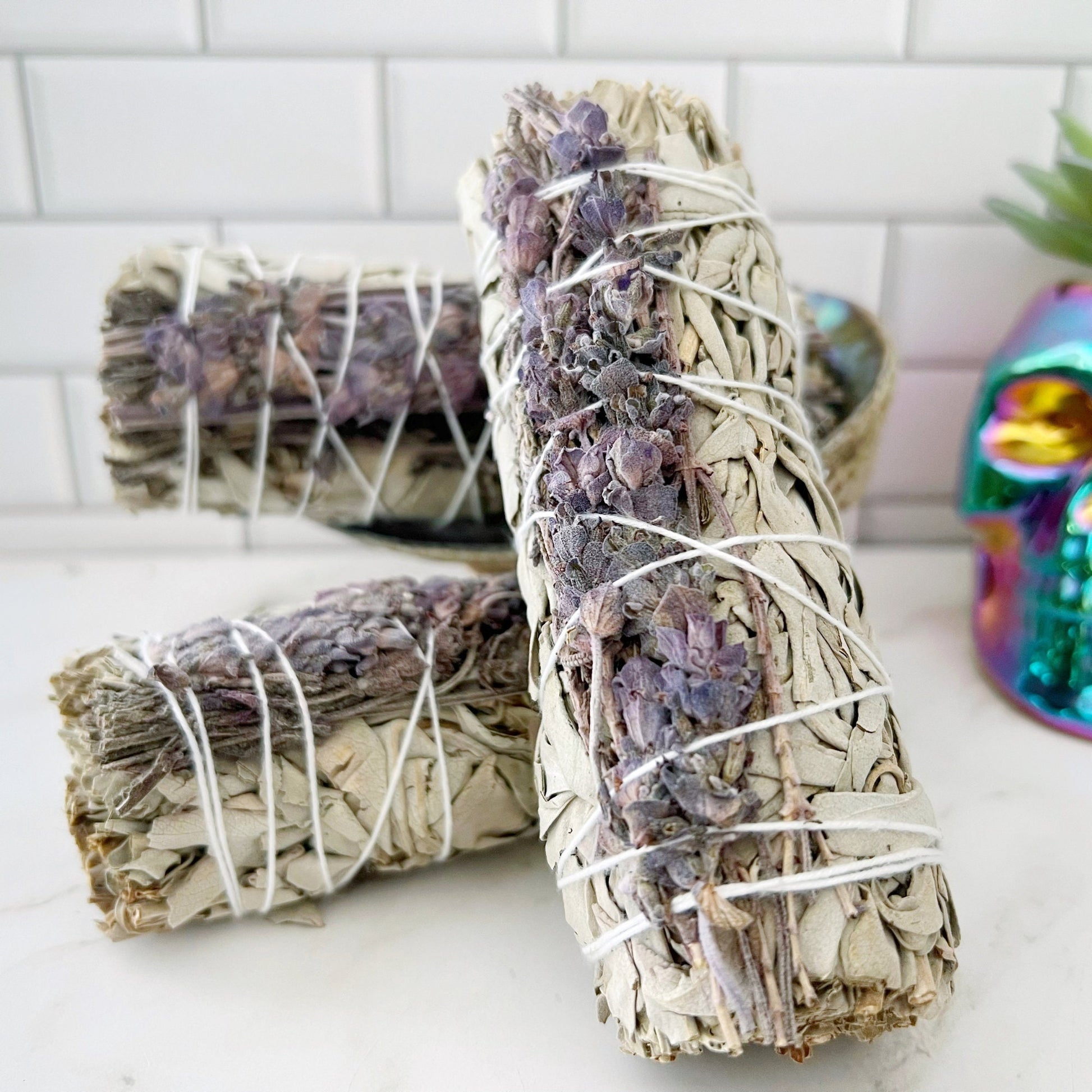 Three **Deluxe Organic White Sage & Lavender Smudge** sticks are bound with string and adorned with dried purple flowers. They rest on a white countertop against a tiled backsplash. In the background, a colorful skull ornament and a small potted succulent add charm to the serene scene.