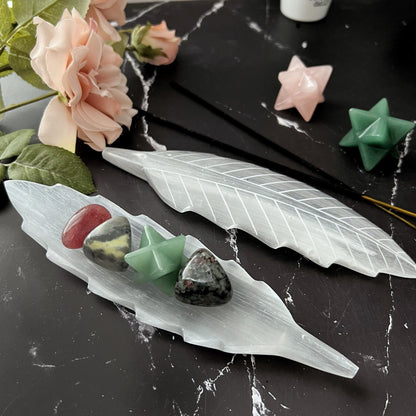 A decorative setup on a black marble surface features a Selenite Feather Incense Holder & Charging Plate elegantly positioned amidst polished stones and star-shaped crystals. Pink roses and green leaves in the background enhance this handcrafted decor, creating a high-vibration aesthetic.