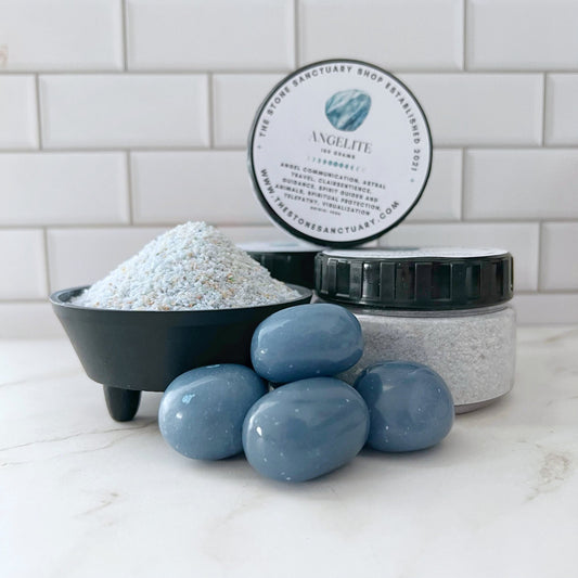 A jar of Angelite Gemstone Sand is displayed behind a small black bowl filled with the scrub and next to a pile of four smooth, blue gemstones that symbolize spiritual connection. The scene is set against a white tiled backsplash and a white countertop.