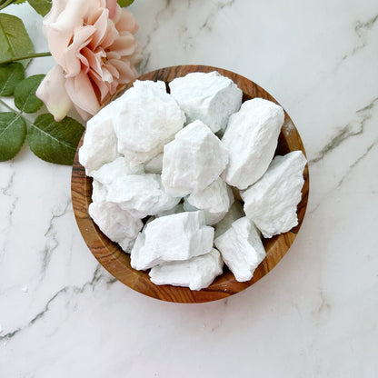 A wooden bowl filled with large, white rock salt chunks and Raw Petalite Stones sits on a white marble surface. A pink rose with green leaves is placed next to the bowl, enhancing the clean and minimalistic setting while inviting a sense of spiritual connection.