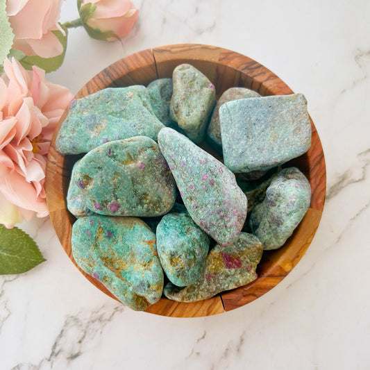 A wooden bowl filled with Raw Ruby Fuchsite Stones, showcasing rough turquoise and green hues with hints of pink and purple speckles. The bowl is set on a white marble surface with a soft pink rose placed beside it, highlighting its UV-reactive properties and metaphysical allure.