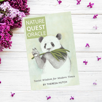 A book titled "Nature Quest Oracle" by Theresa Hutch rests on a white wooden surface surrounded by purple flowers. The cover features an illustrated panda holding a bamboo leaf, inviting readers to explore Taoist philosophy for a more mindful life.