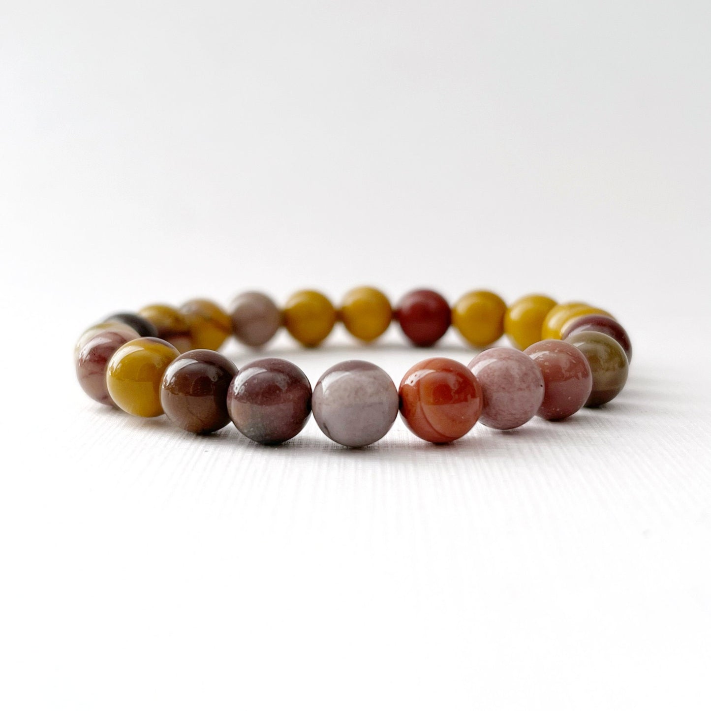 A 8mm Mookaite Beaded Bracelet made of multicolored polished stones is displayed on a white surface. Behind the gemstone bracelet, there is a golden bowl containing more beads, and a pink rose in soft focus on the left side adds a gentle, elegant touch to the scene.