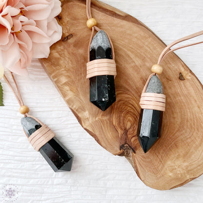 Three Black Tourmaline Leather Wrapped Point Necklaces with wooden beads are arranged on a wooden surface. Pink flowers are placed beside the crystals, creating an elegant and natural aesthetic of protective stone jewelry.