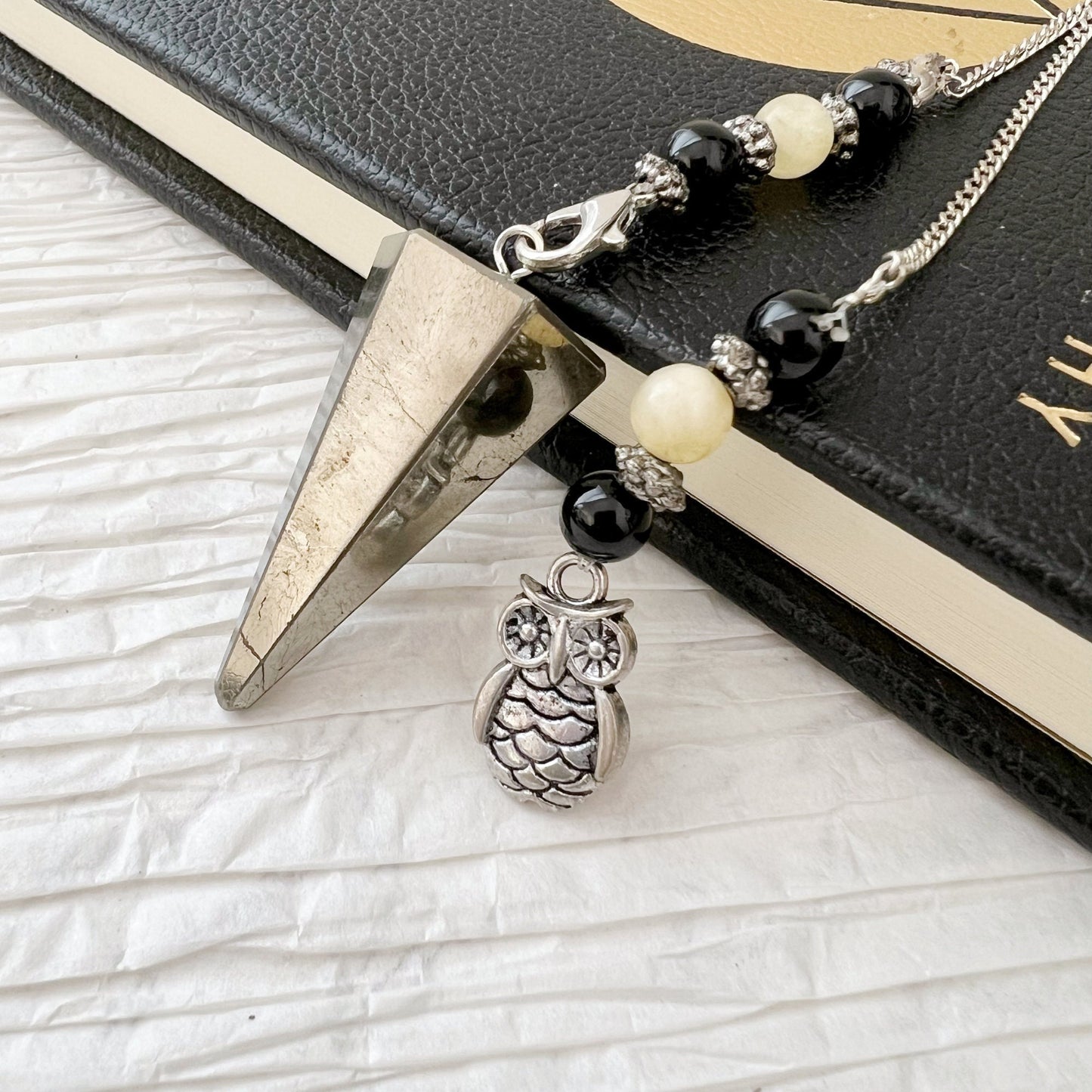 A Pyrite Hexagonal Pendulum with Owl Charm and a beaded bracelet featuring onyx beadwork with black and white beads are draped over a black and gold book. The bracelet boasts a silver owl charm hanging from it. The background is a white textured surface.