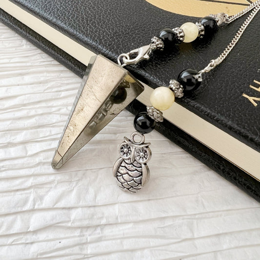 A Pyrite Hexagonal Pendulum with Owl Charm and a beaded bracelet featuring onyx beadwork with black and white beads are draped over a black and gold book. The bracelet boasts a silver owl charm hanging from it. The background is a white textured surface.