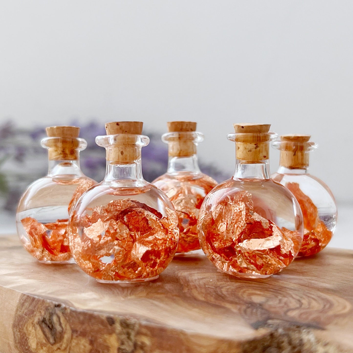 Six Copper Flakes Bottles with cork stoppers are filled with shimmering copper flakes, ideal for chakra healing and other metaphysical practices. The bottles are arranged on a wooden surface with a blurred background, highlighting the intricate texture and vibrant color of the copper inside the clear jars.