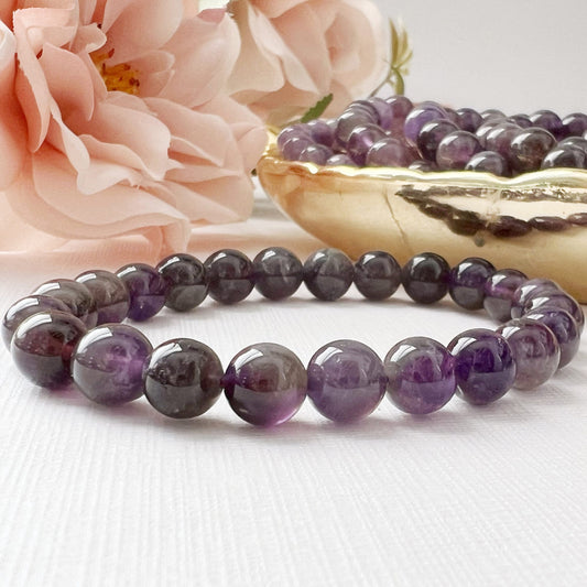 A close-up of the 8mm Amethyst Beaded Bracelet placed on a light surface, with a pink peony flower and a brass bowl filled with more amethyst beads in the background. The meditation jewelry features smooth, polished beads displaying various shades of purple.