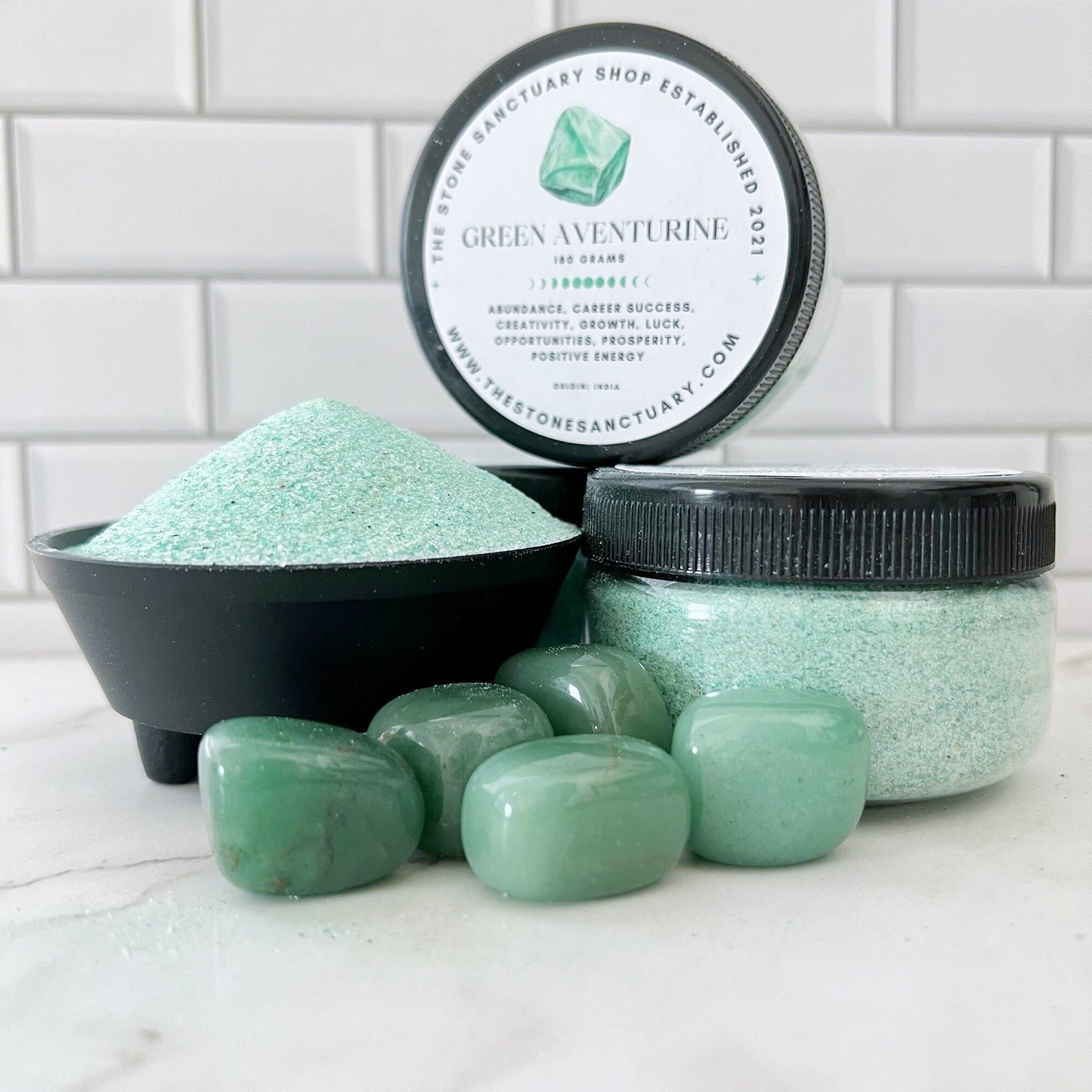 A set of Green Aventurine Gemstone Sand items includes a jar and a black bowl of powdered aventurine, along with seven polished green aventurine stones. The backdrop is a white tiled wall, and the jar is labeled with information about Green Aventurine Gemstone Sand's benefits for luck and prosperity.