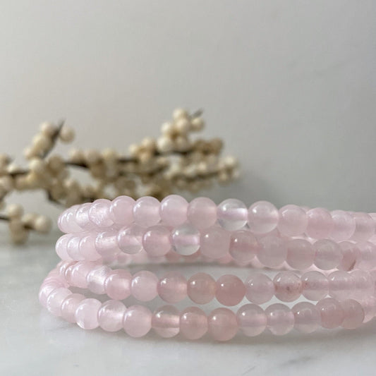 A close-up image shows a 4mm Rose Quartz Beaded Bracelet made of semi-precious stones, coiled in a circular pattern. The backdrop features an out-of-focus branch adorned with small white berries, set against a light, soft-focus background, enhancing the bracelet’s connection to Heart Chakra healing.