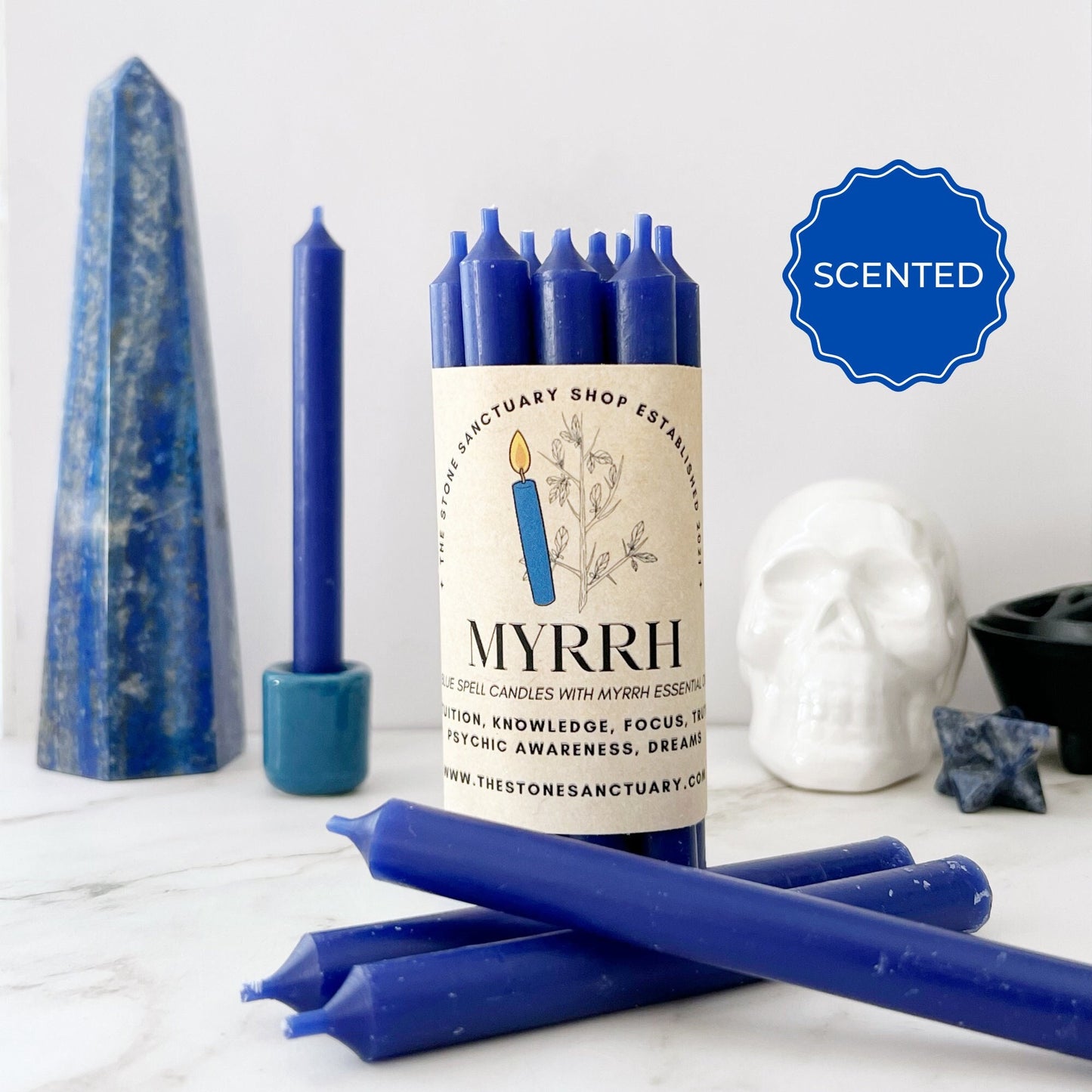 A collection of 5" Myrrh Scented Blue Chime Candles is displayed, with one in a cylinder wrapping and several individual candles lying around. The background includes a tall blue crystal, a white skull decoration, a dark circular object, and a blue star-shaped stone.