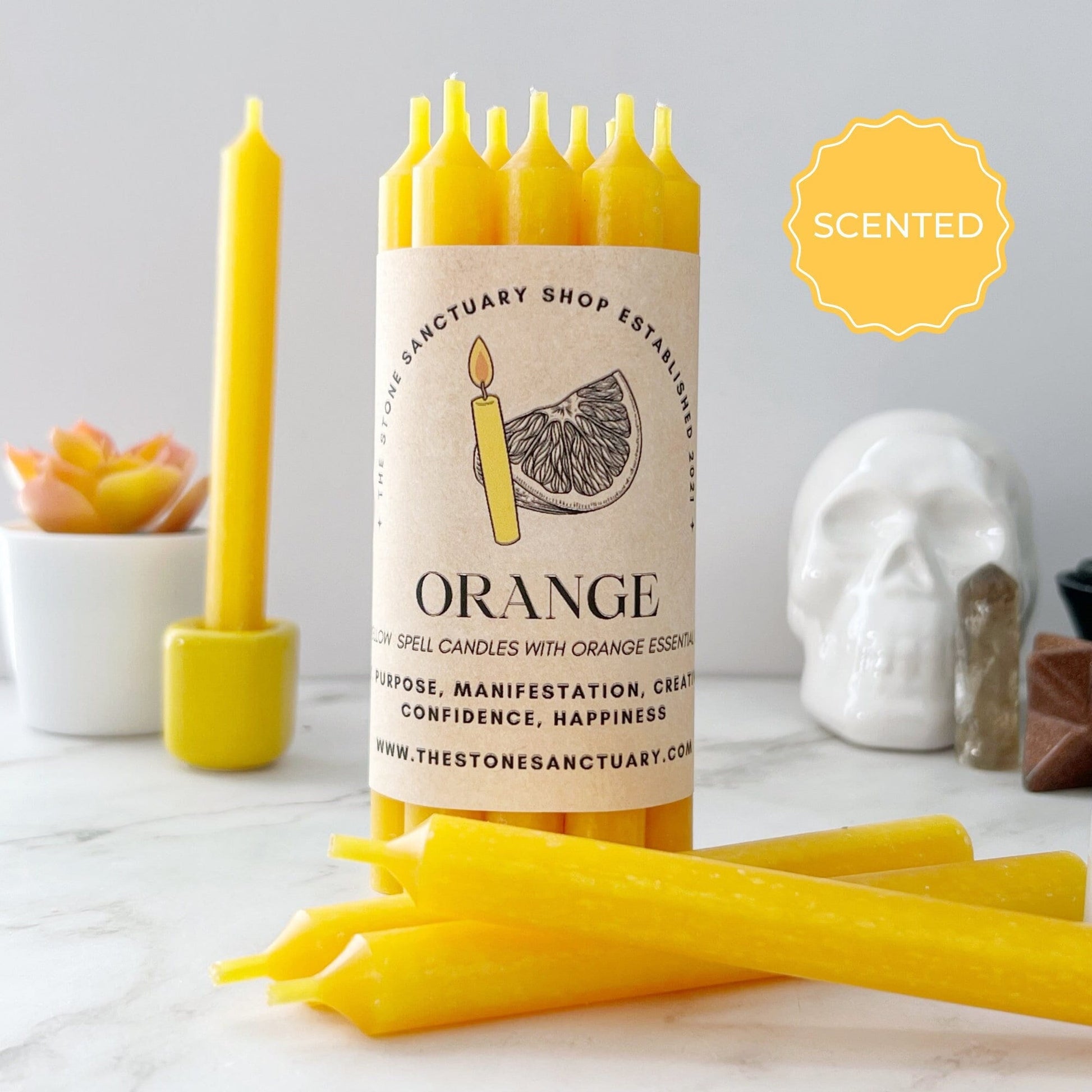 A display of 5" Orange Scented Yellow Chime Candles labeled "Orange" with a tag noting "Purpose, Manifestation, Creativity, Confidence, Happiness." A succulent and skull decoration appear in the background. Featuring orange oil for a fragrant touch, these candles are perfect for igniting your creative spirit.