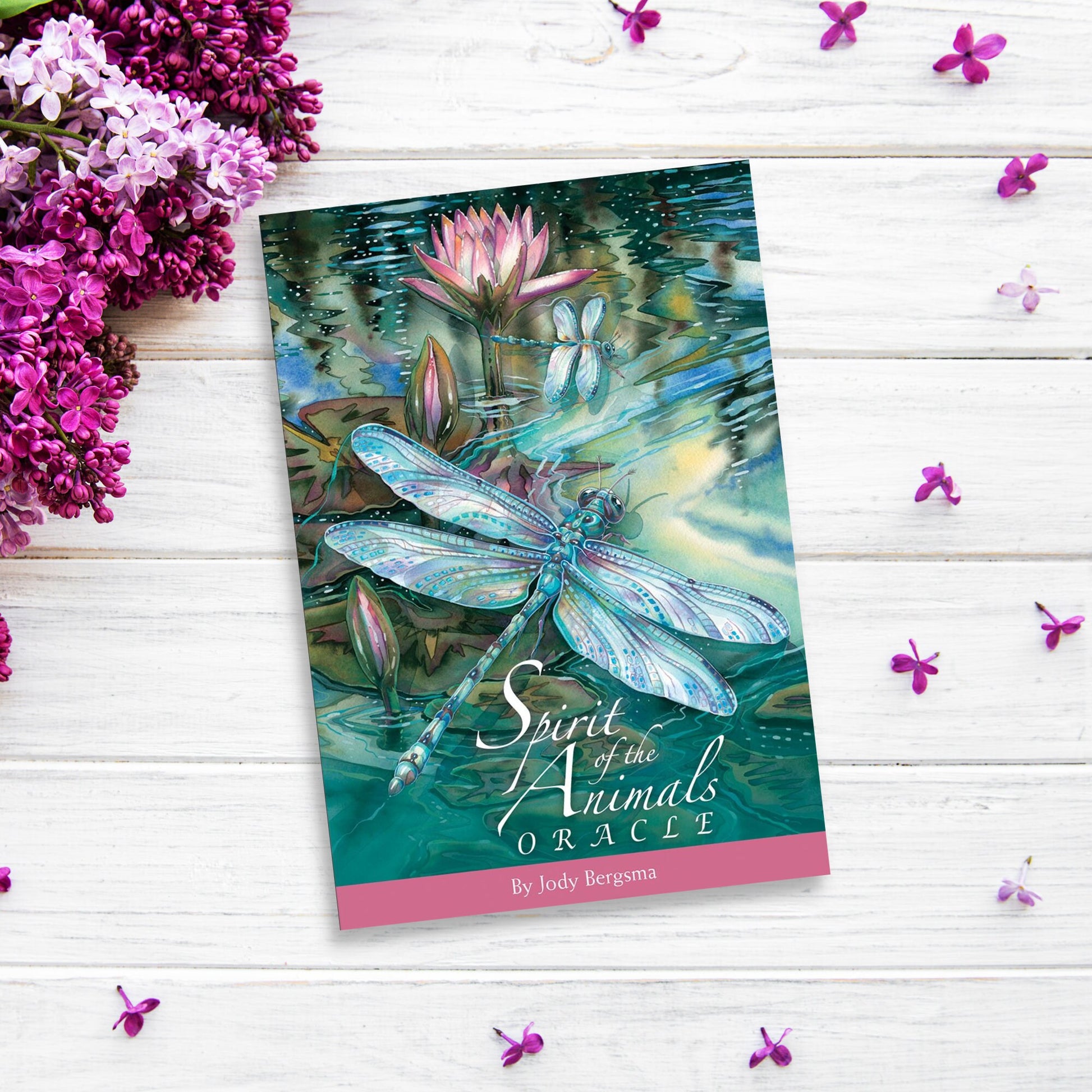 A card deck titled "Spirit Of The Animals Oracle" by Jody Bergsma is laid on a white wooden surface. The cover features a dragonfly and water lilies, embodying the essence of Animal Spirit Cards. Surrounding the deck are purple flowers and small purple petals scattered on the surface.