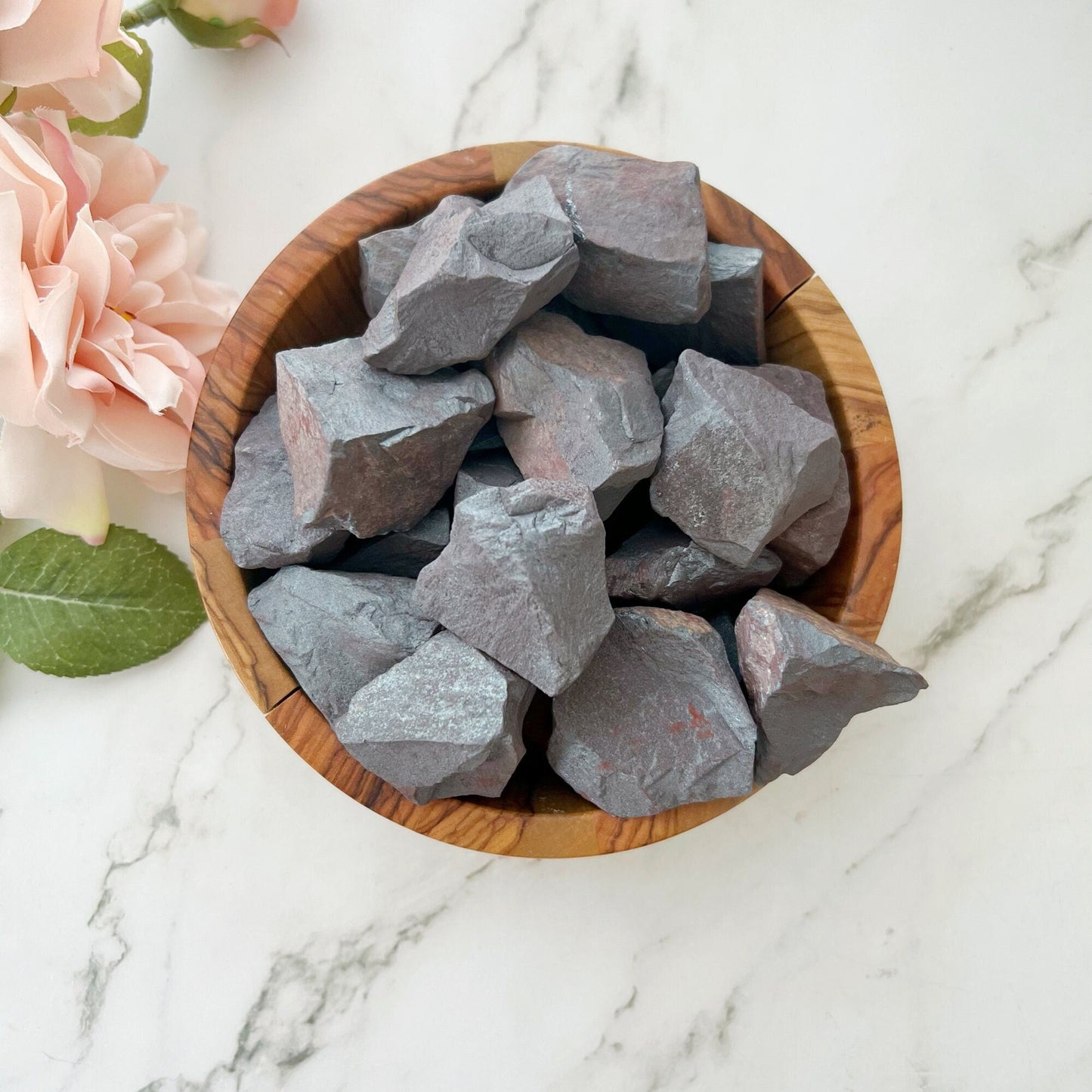 A wooden bowl filled with rough, dark grey Raw Hematite Stones sits on a white marble surface. The bowl is surrounded by light pink roses with green leaves, adding a touch of color to the scene—perfect for grounding yourself and embracing the protective properties of these crystals.