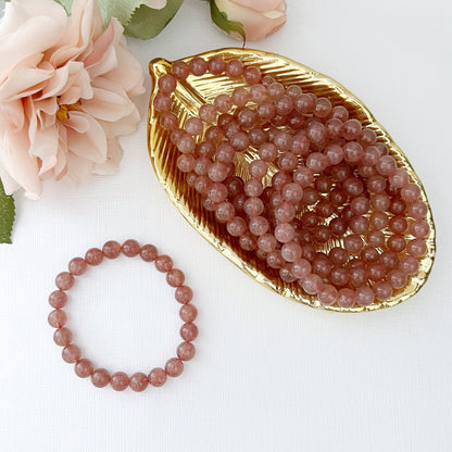 An 8mm Strawberry Quartz Beaded Bracelet made of spherical, peach-colored beads rests on a white surface. In the background, there are peach-colored roses slightly blurred, and a golden dish filled with similar beads. The overall setting is elegant and softly lit.