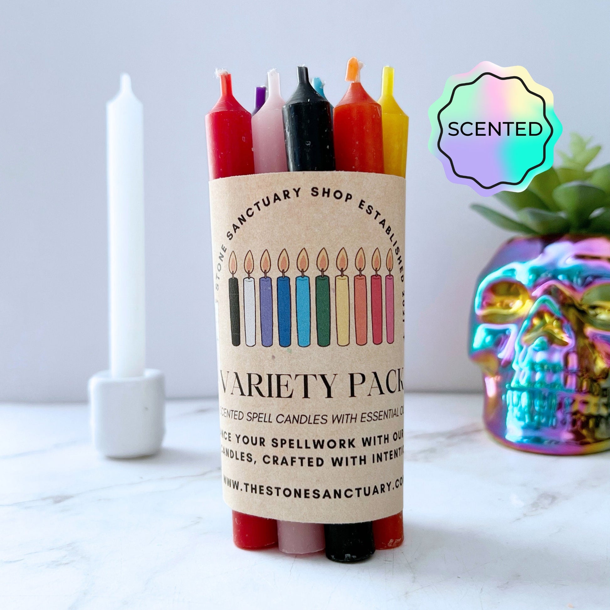 A Variety Pack Scented Chime Candles is displayed on a marble surface. The pack contains six different colored candles, each aligned with chakra balancing, and showcases a label with multiple candle illustrations. Nearby, a tall white candle is visible, along with a colorful, reflective skull decoration.