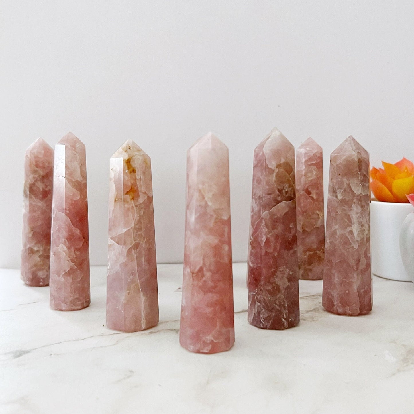 A photo featuring seven 3-4" Rose Quartz Six Sided Standing Points arranged in a semi-circle showcases their natural marbling and varying shades of pink. These stones are not just beautiful but also serve as a powerful crystal energy generator for emotional healing. In the background, a small pot with a yellow-orange succulent is partially visible on the light-colored surface.