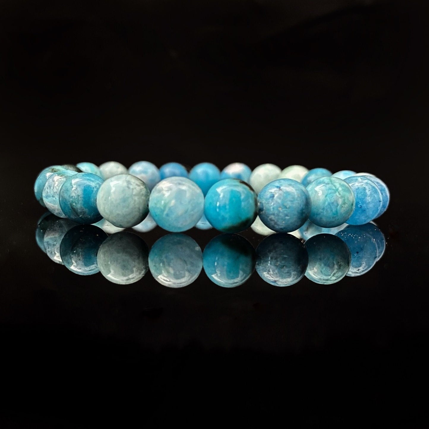 A stunning 8mm Hemimorphite Beaded Bracelet made of round, polished blue stones is displayed against a black background. The stones vary in shades of blue and exhibit slight marbling, emanating Reiki energy. The bracelet's reflection shimmers on the shiny surface beneath it.