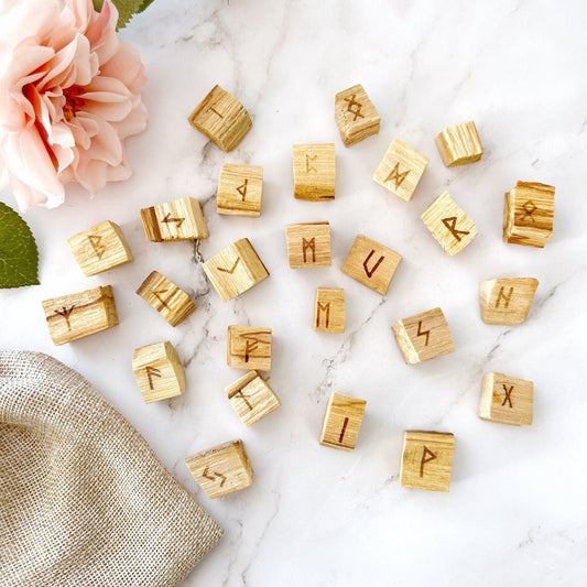 A Palo Santo Wood Runes, featuring the ancient Elder Futhark symbols, is displayed on a marble surface. A pink flower and a piece of burlap cloth are placed next to the blocks, adding a touch of natural decoration and highlighting their sustainable harvesting background.