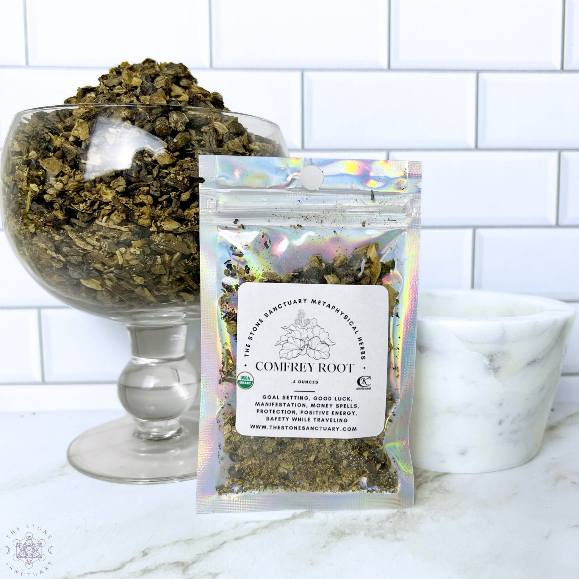 A holographic pouch of Comfrey Root from The Stone Sanctuary sits in front of a large, overflowing glass bowl of dried comfrey root. A white mortar and pestle are on the right, perfect for preparing ingredients for magical rituals or money spells. The background features white subway tile.