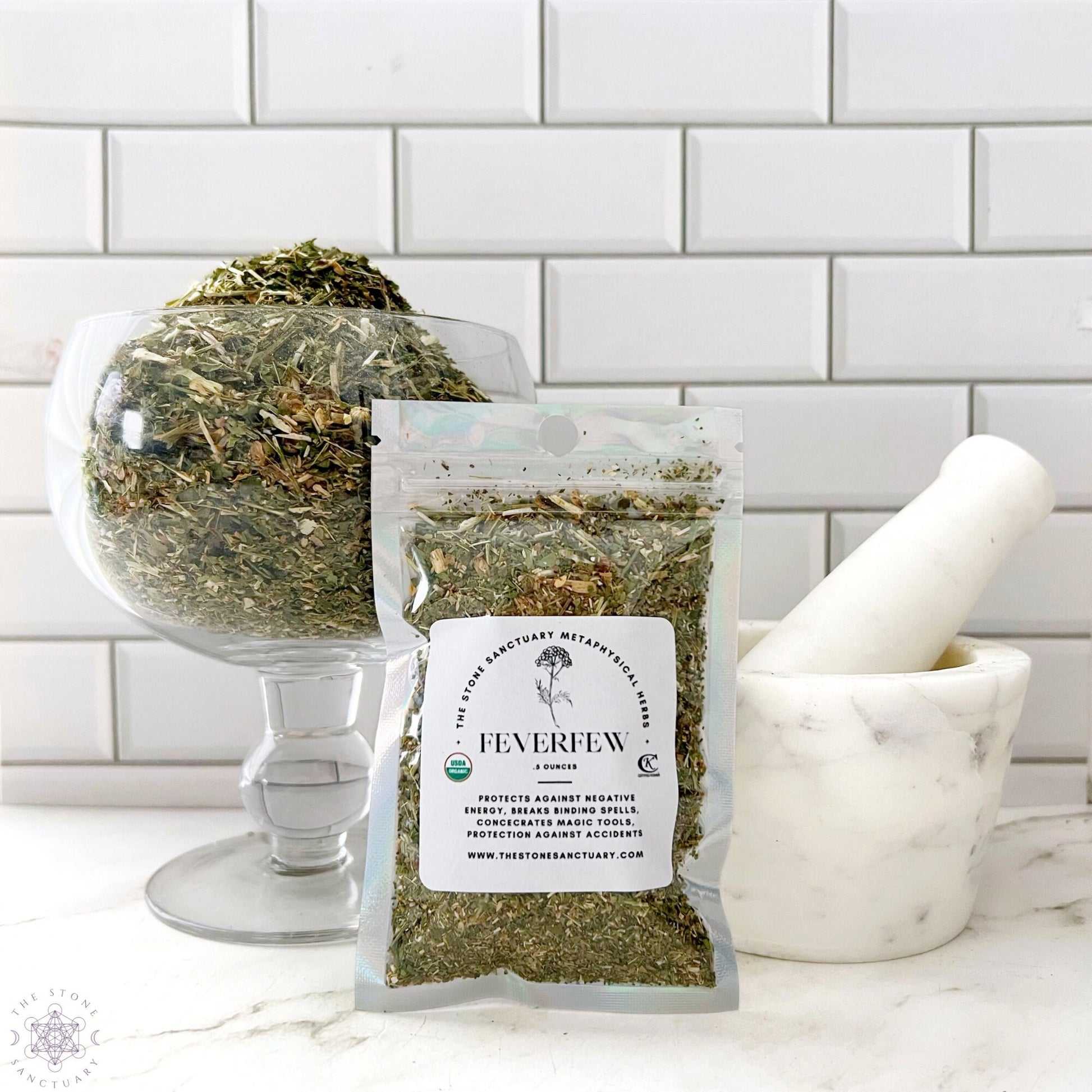 A clear glass bowl filled with dried feverfew herb is next to a small white marble mortar and pestle. In front of the bowl, there is a sealed plastic bag labeled "Feverfew Leaves" with descriptions and certifications. The background features white tiled walls.