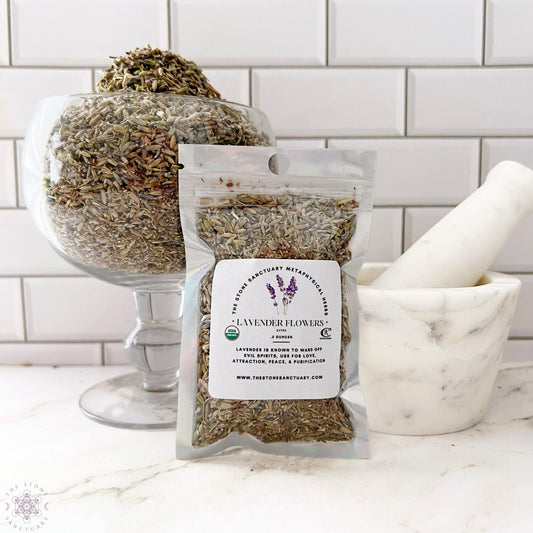 A sealed bag of Lavender Flowers stands in front of a glass bowl filled with more dried lavender, perfect for relaxation and calmness. To the right, a white marble mortar and pestle rest on a white countertop with a tiled backsplash. The bag's label lists the product details.