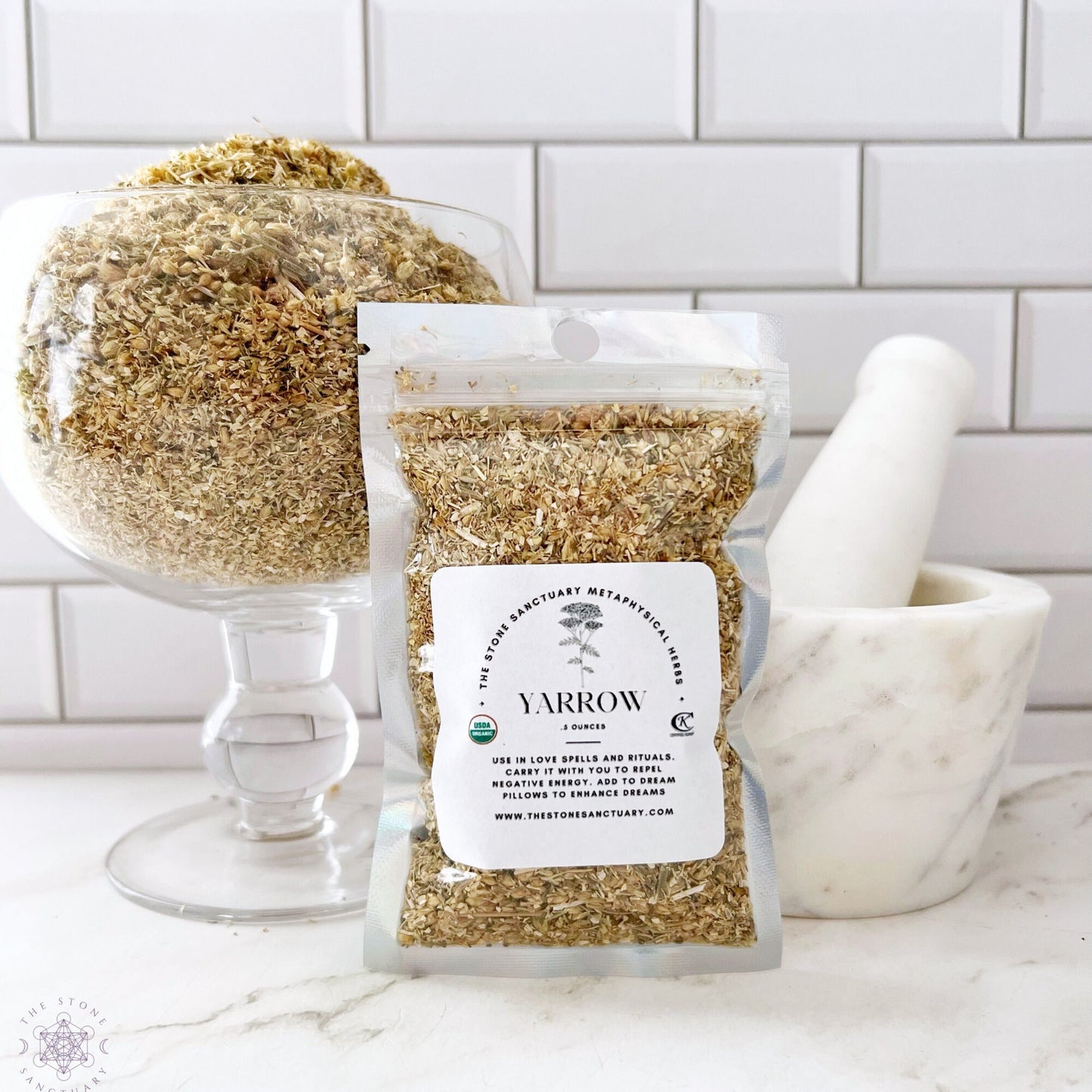 A small sealed package of Yarrow Flower sits on a counter with loose yarrow filling a stemmed glass container in the background. To the right, a white mortar and pestle are visible. The backdrop consists of a white tile wall, perfect for spell casting or enhancing your psychic powers.