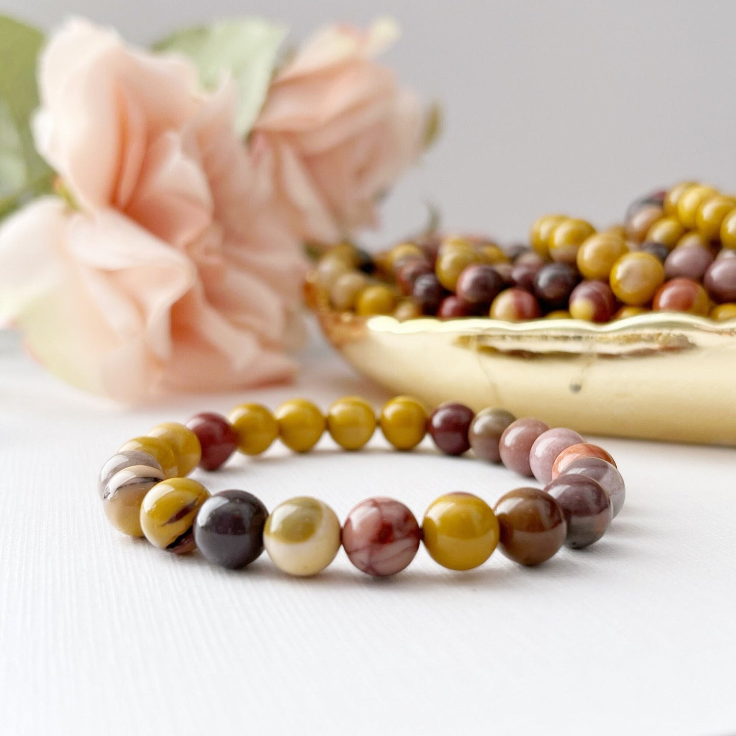 A 8mm Mookaite Beaded Bracelet made of multicolored polished stones is displayed on a white surface. Behind the gemstone bracelet, there is a golden bowl containing more beads, and a pink rose in soft focus on the left side adds a gentle, elegant touch to the scene.