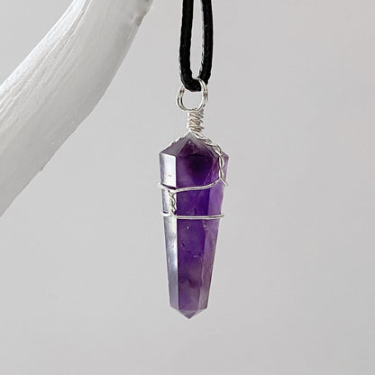 A close-up image of a 1.5" Double Terminated Amethyst Point Pendant featuring a polished, double-terminated stone wrapped with thin silver wire. Known for promoting emotional balance, the pendant is suspended from a black cord and displayed against a plain gray background.