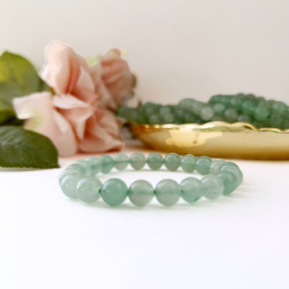 An 8mm Green Aventurine Beaded Bracelet, imbued with calming energy, rests on a white surface in front of a pink rose flower. In the background, a brass dish holds another similar 8mm Green Aventurine Beaded Bracelet, slightly out of focus. The overall composition gives a soft, elegant appearance.